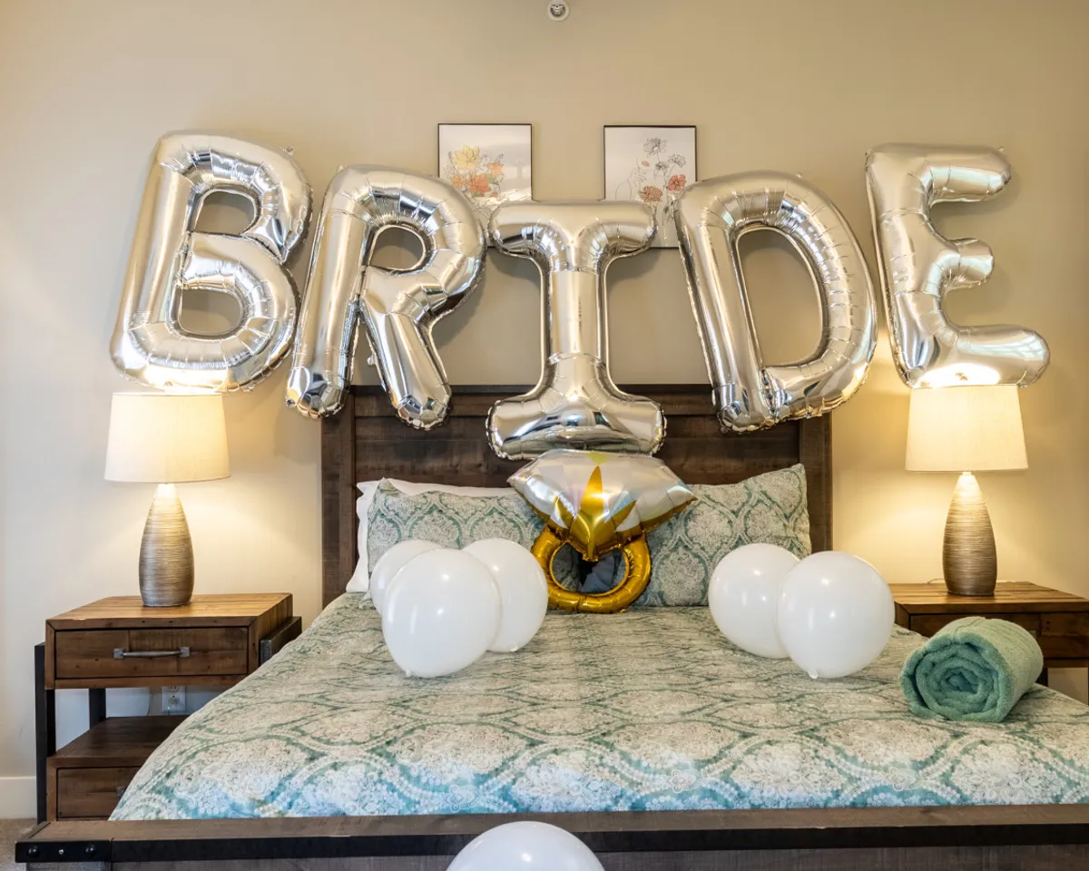 A cheerful bedroom scene with balloon letters spelling 'BRIDE,' decorative pillows, white and gold balloons, and soft bedside lighting. The celebratory decor signals the space's suitability for group gatherings or special occasions.