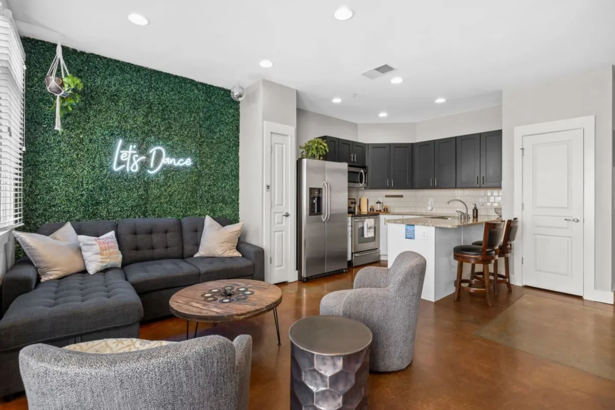 A lively open-plan area where a chic sofa and coffee table face a kitchen in the background. The green wall with a 'Let's Dance' neon sign adds a playful, inviting touch, while the kitchen area is visible and ready for entertaining or culinary adventures. The overall vibe is cheerful, energetic, and perfect for gatherings.