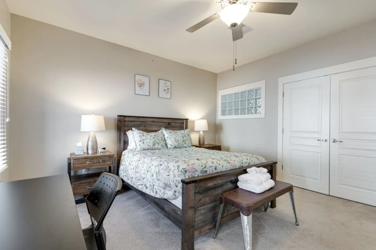 A tranquil upstairs bedroom scene featuring a queen-size bed with floral bedding, flanked by nightstands and lamps, and complemented by a desk and chair. The room is bathed in natural light, creating a serene retreat ideal for rest, relaxation, or quiet work.