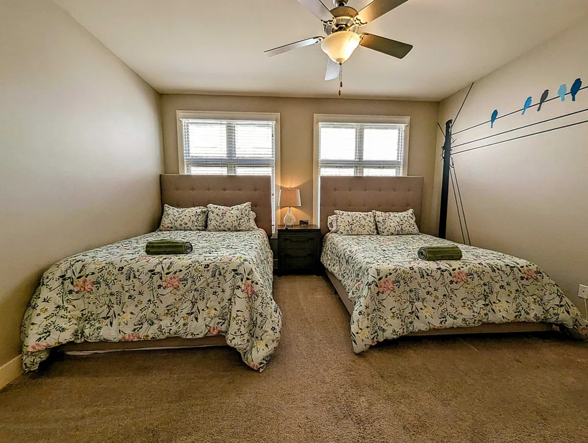 A serene bedroom featuring two beds with floral bedding, a nightstand illuminated by a lamp, and decorative wall art of birds above the beds. The room is inviting and filled with natural light, creating a comfortable atmosphere for resting or unwinding after a long day.