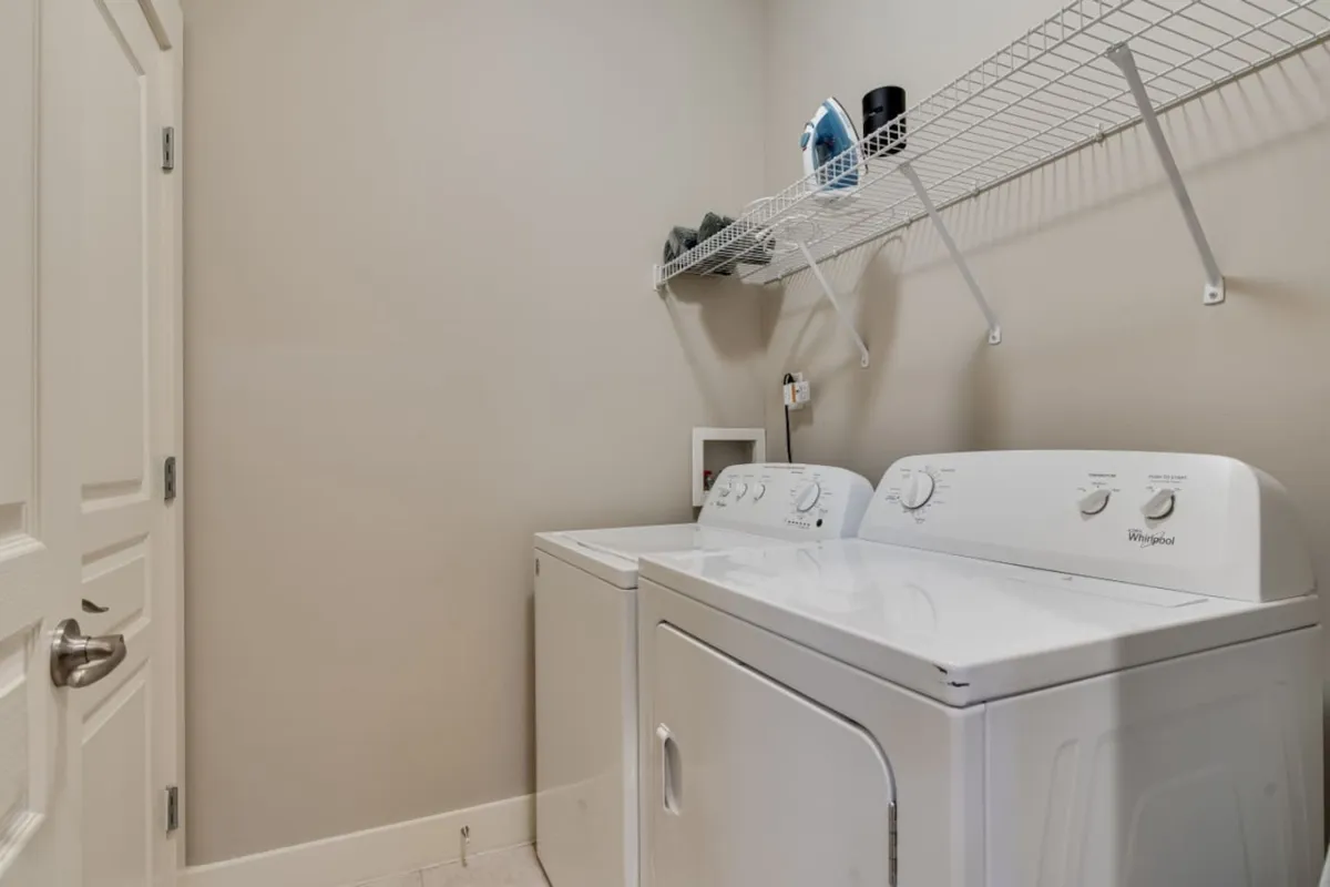 A practical and neatly organized laundry area featuring a washer, dryer, and a storage shelf with an iron. The space is functional and conveniently located, facilitating easy laundry routines during long or short stays.