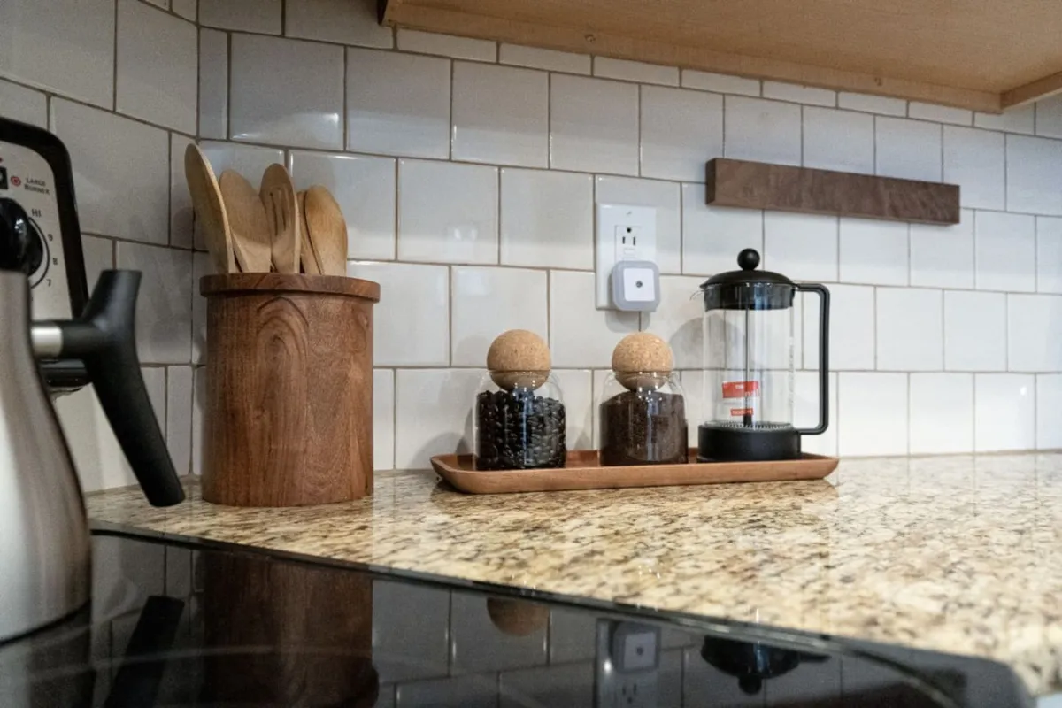 A close-up look at the well-equipped kitchen, showcasing wooden utensils, glass jars filled with coffee beans, a French press, and a stylish granite countertop. The scene suggests both practicality and warmth, inviting residents and guests to brew fresh coffee or prepare meals together.